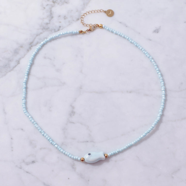 Necklace with light blue beads and a white fish-shaped pendant on a marble surface