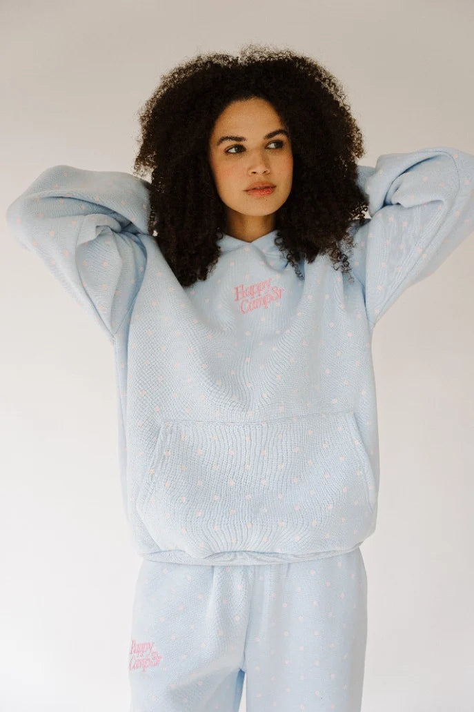 A woman with curly dark hair wearing a light blue textured knit matching loungewear set with a small pink graphic on the chest and thigh, posing against a plain light gray background.