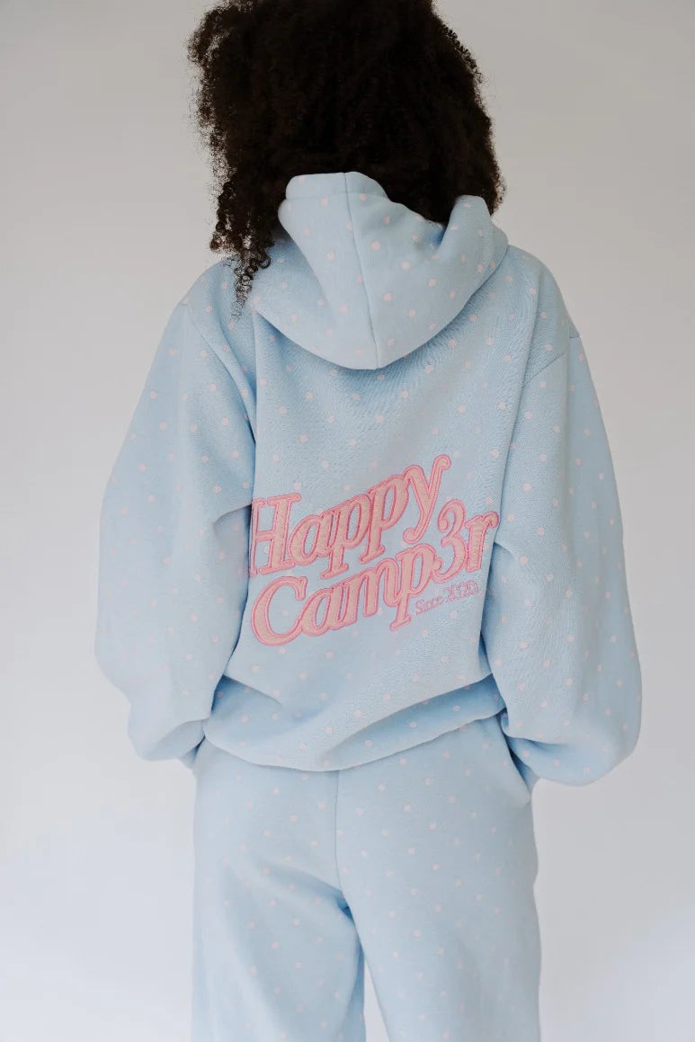 A person shown from the back wearing a light blue hooded sweatshirt and matching sweatpants with small white polka dots. The hoodie has a large pink 'Happy camper' graphic across the back.