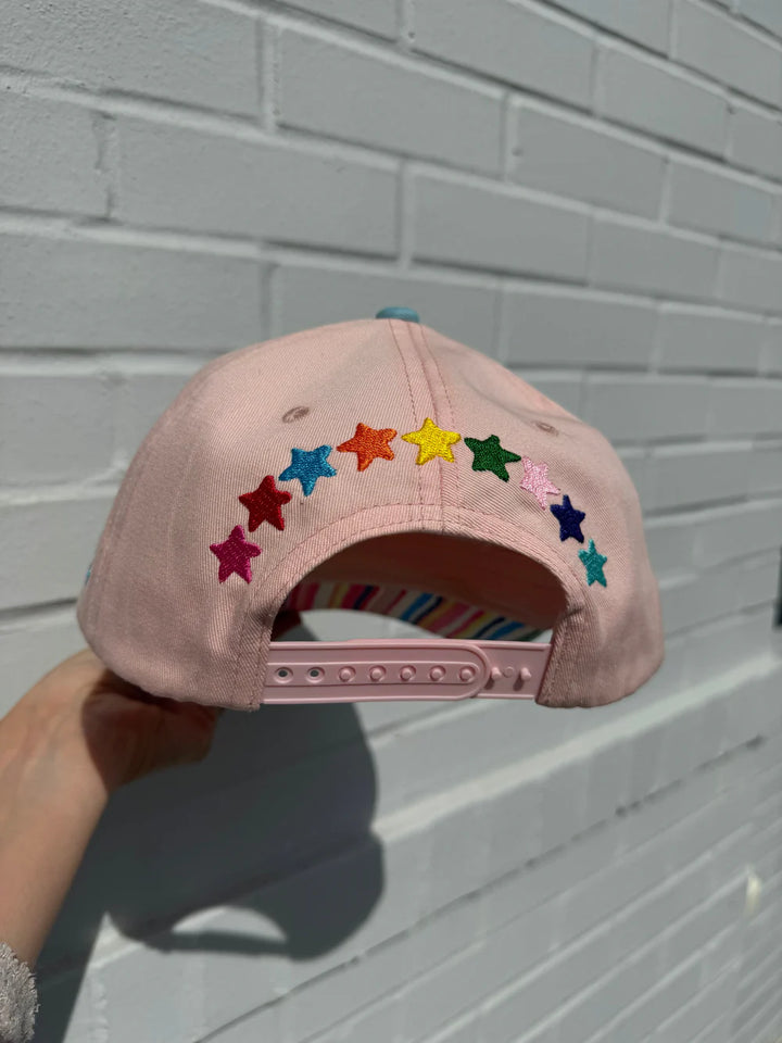 Pink cap with colorful star patterns held against a light gray brick wall.