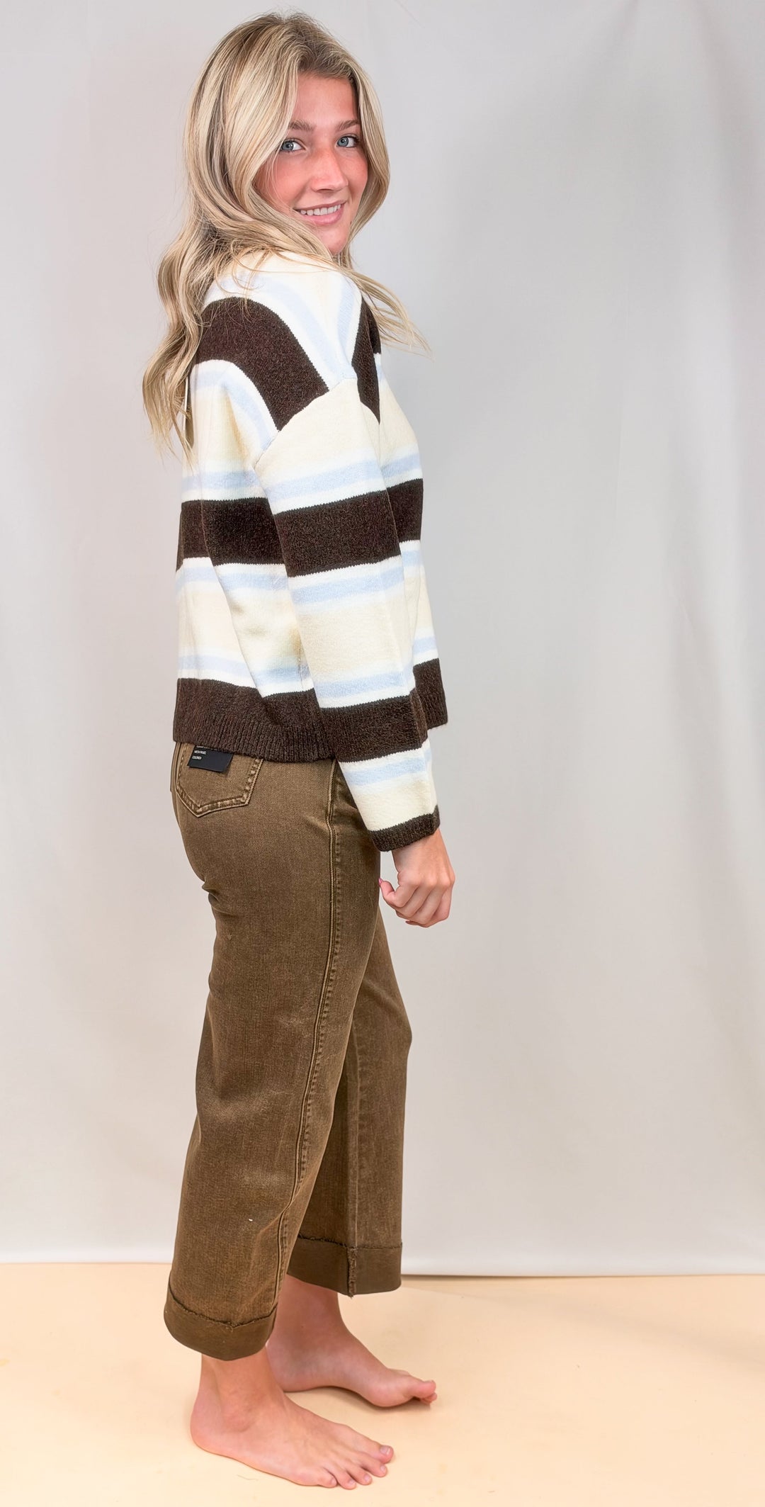 Woman shown from side view wearing striped cardigan with brown jeans on plain background