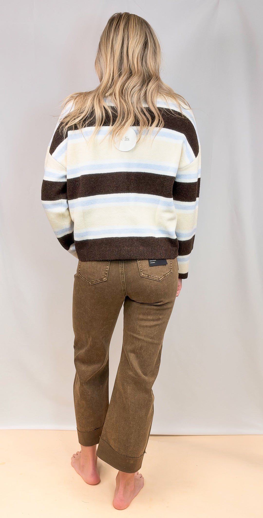 Woman shown from back view wearing striped cardigan and brown jeans on plain background