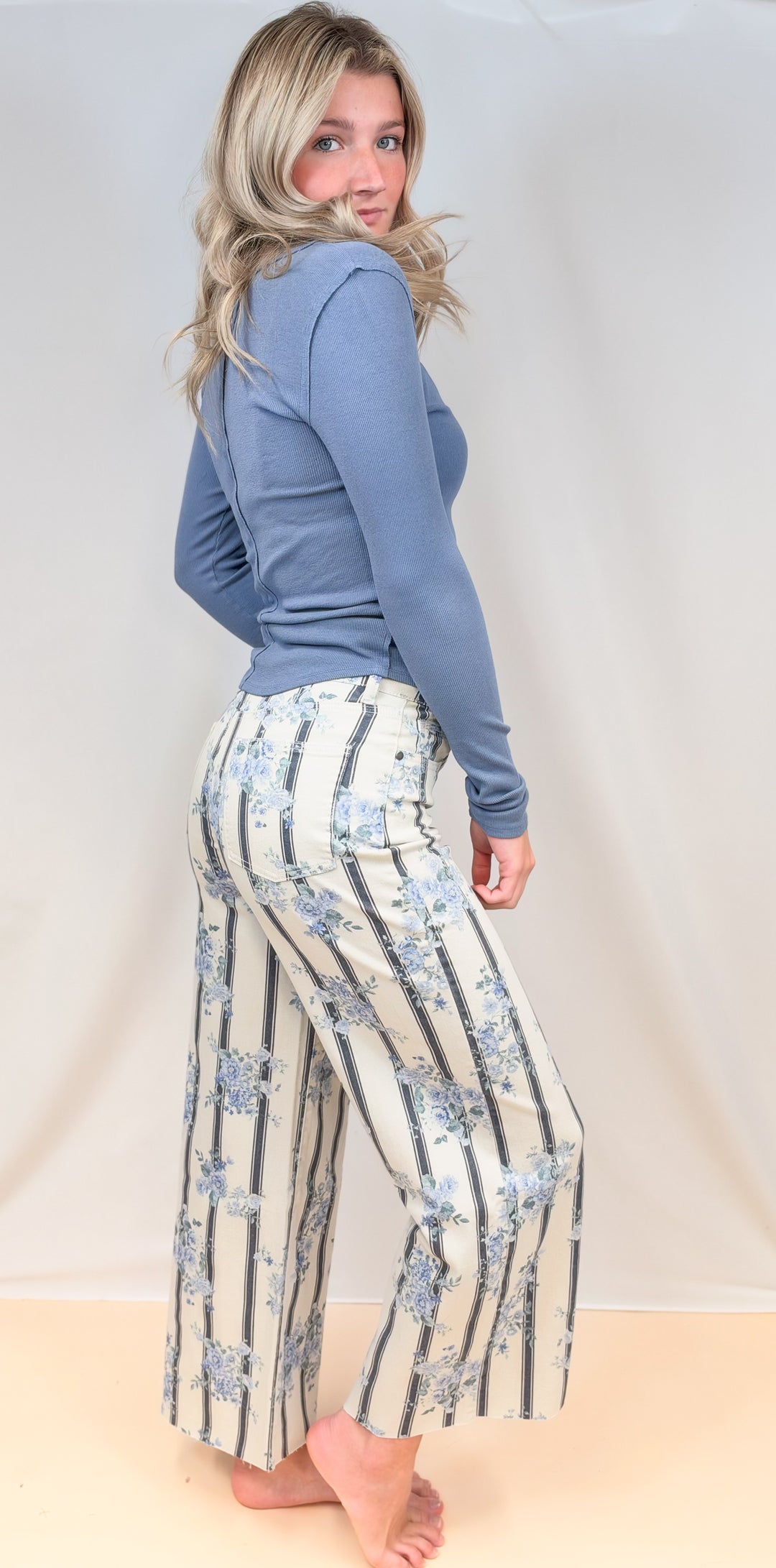 Woman shown from side view wearing blue long sleeve top with white and blue stripe pants with floral detail on plain background