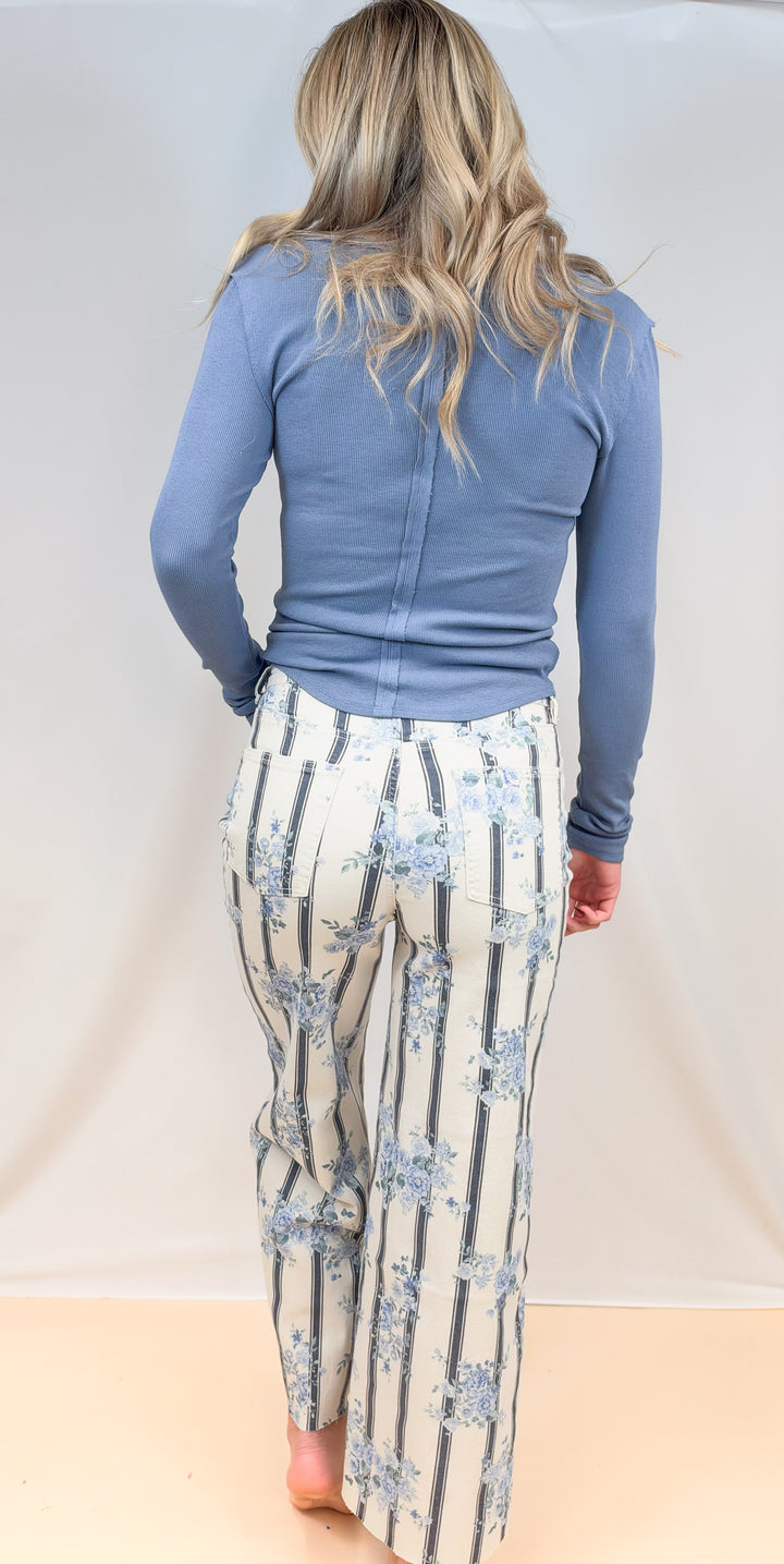 Woman shown from back view wearing blue long sleeve top with white and blue stripe pants with floral detail on plain background