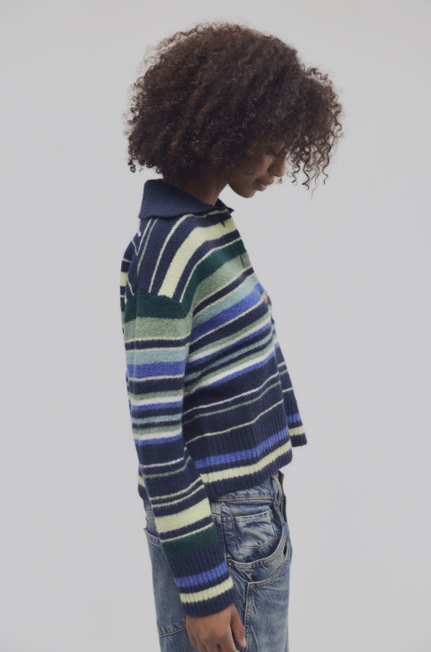 A model with curly dark hair is shown from a side profile wearing a striped knit polo sweater with navy, blue, green, and cream stripes, paired with blue jeans, against a solid light gray background.