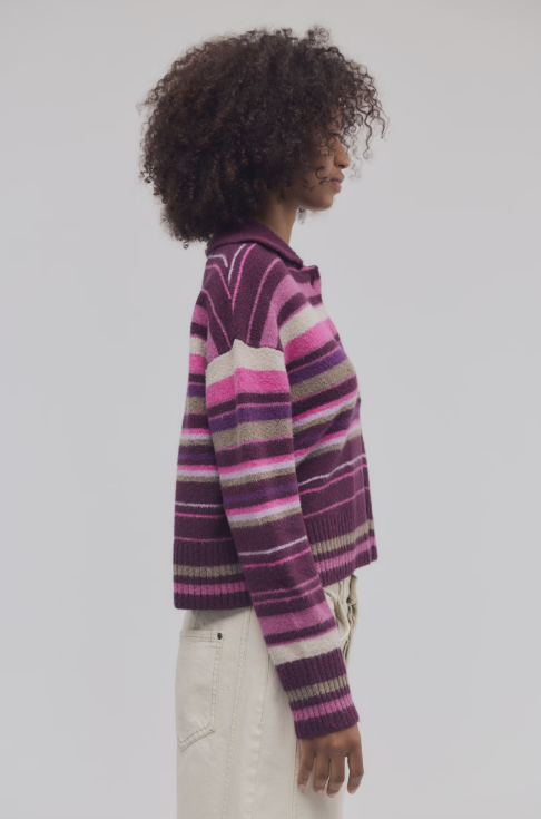 A woman shown in side profile wearing a cropped knit sweater with horizontal stripes in purple, pink, and beige, paired with off-white pants, against a plain light gray studio background.
