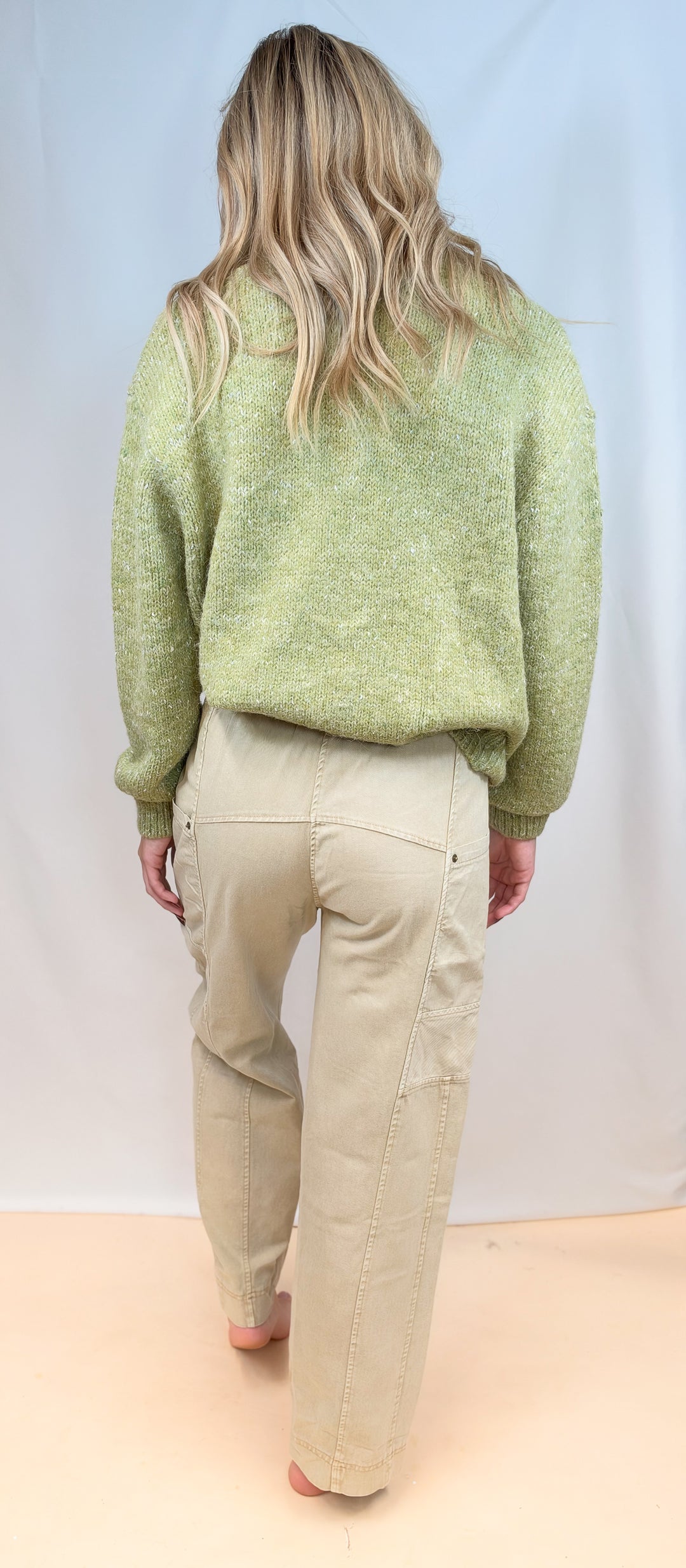 Woman shown from back view wearing green sweater and beige pants on plain background