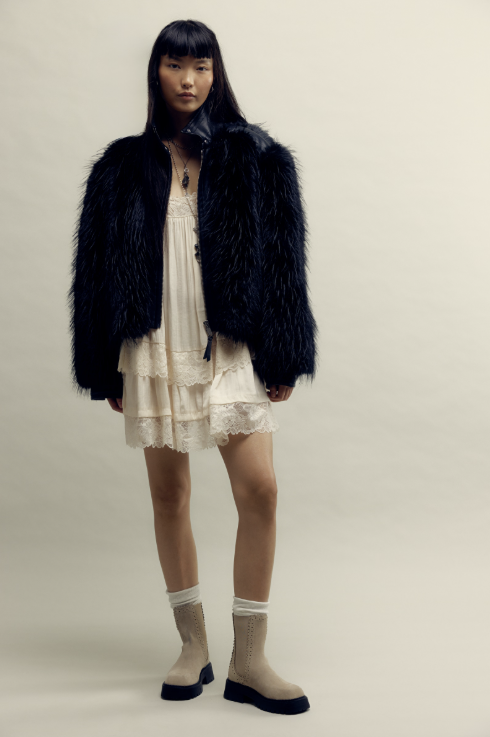 A model stands against a plain light beige studio background wearing a black faux-fur jacket over a cream lace dress, with beige socks and black platform boots.