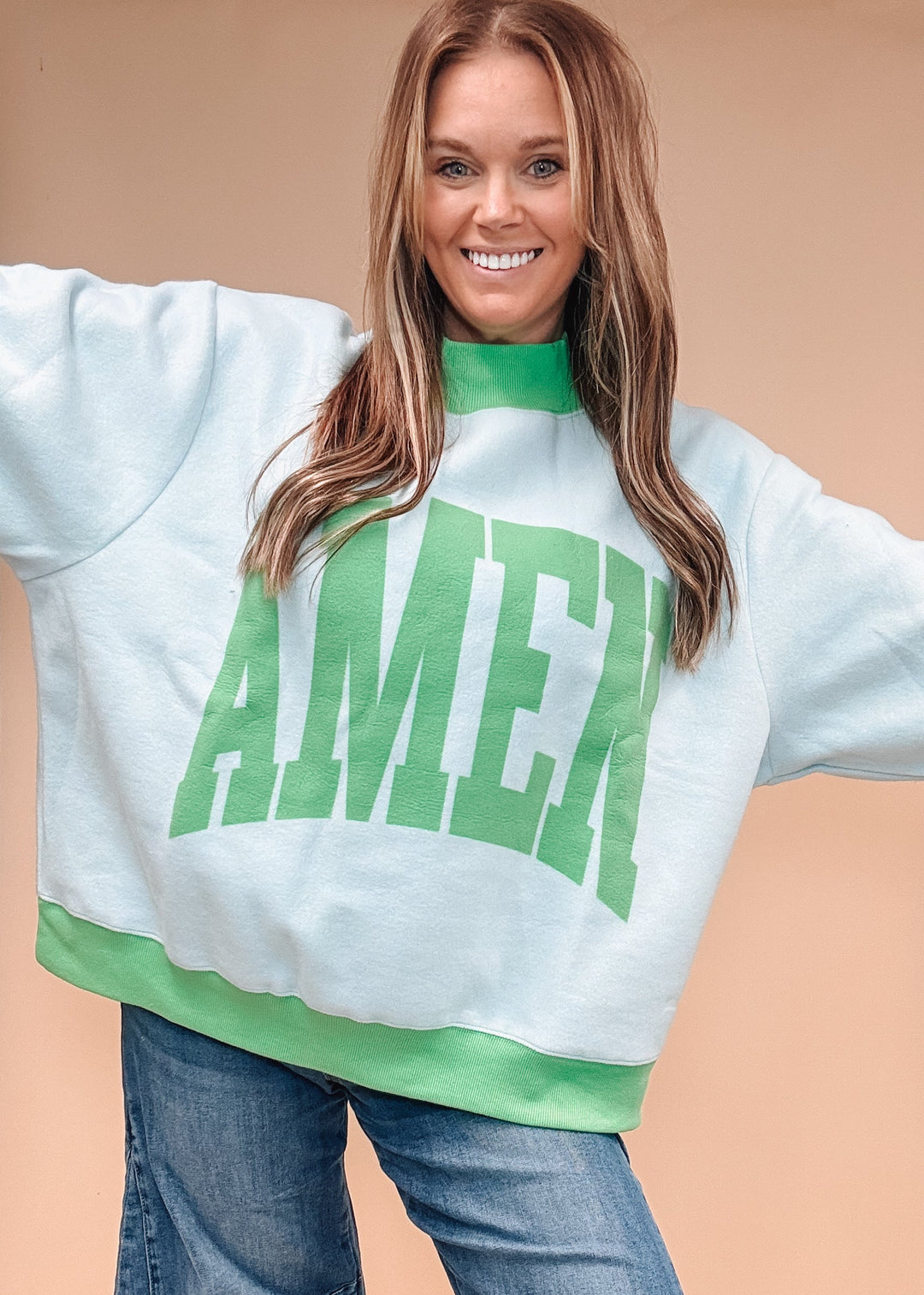 A woman wearing a light blue sweatshirt with a green mock neck, green ribbed hem, and a large green &