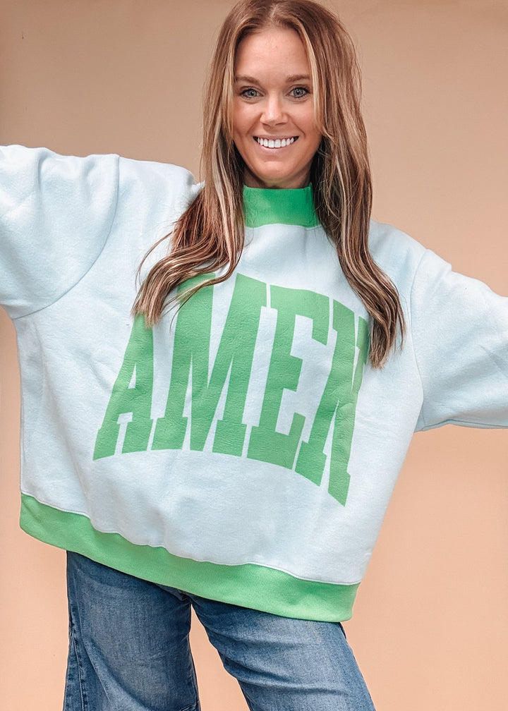 A woman wearing a light blue sweatshirt with a green mock neck, green ribbed hem, and a large green &