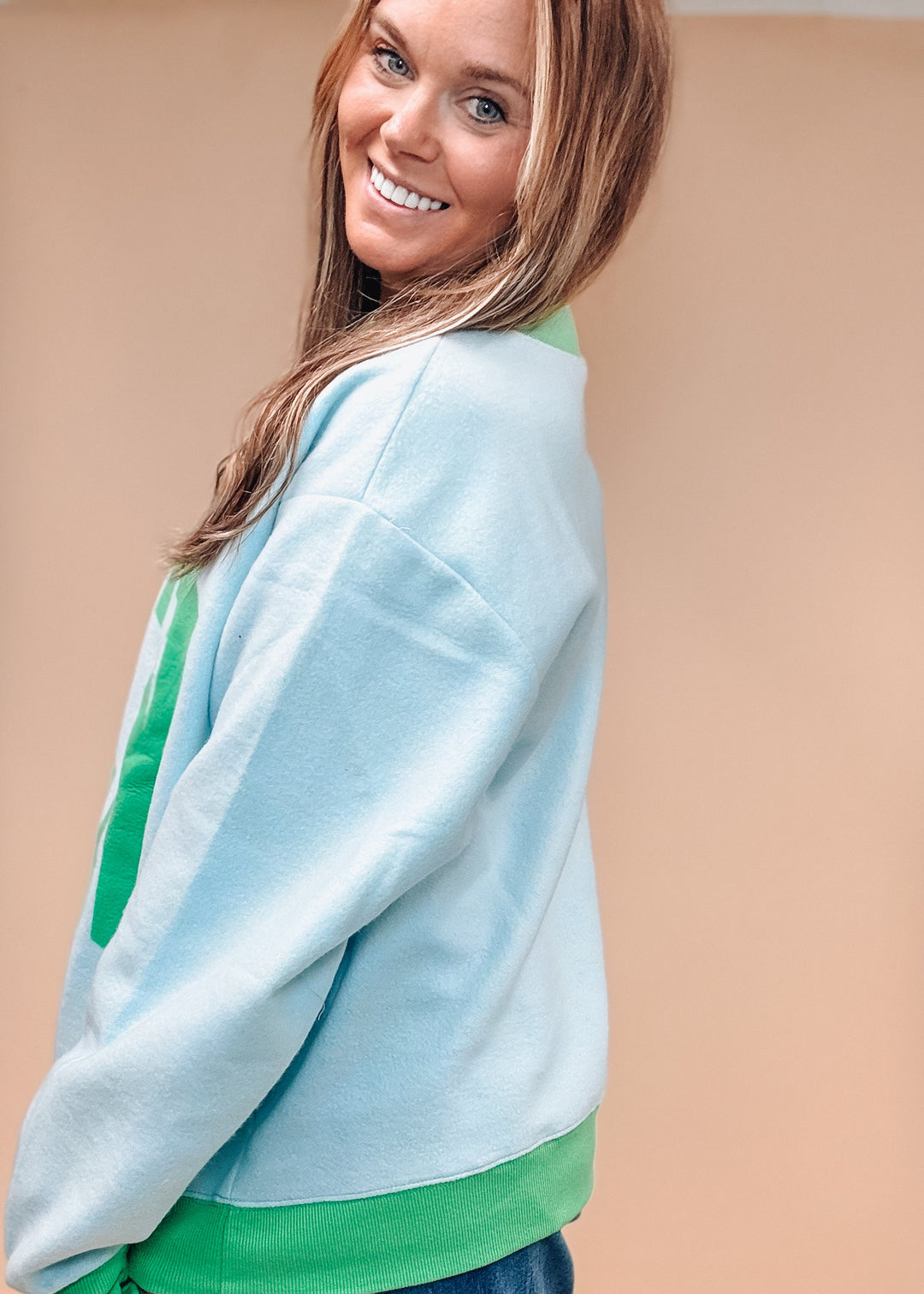 A woman wearing a light blue and green color-blocked sweatshirt with blue jeans, photographed against a solid beige background.