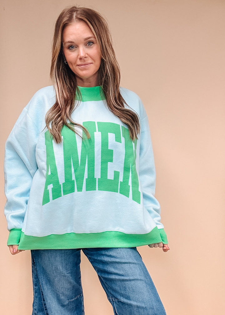 A woman modeling a light blue crewneck sweatshirt with green trim and a large green &