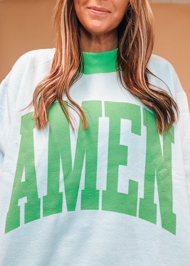A close-up of a woman wearing a white crewneck sweatshirt with a green mock neck, green ribbed hem and cuffs, and a large green collegiate-style &