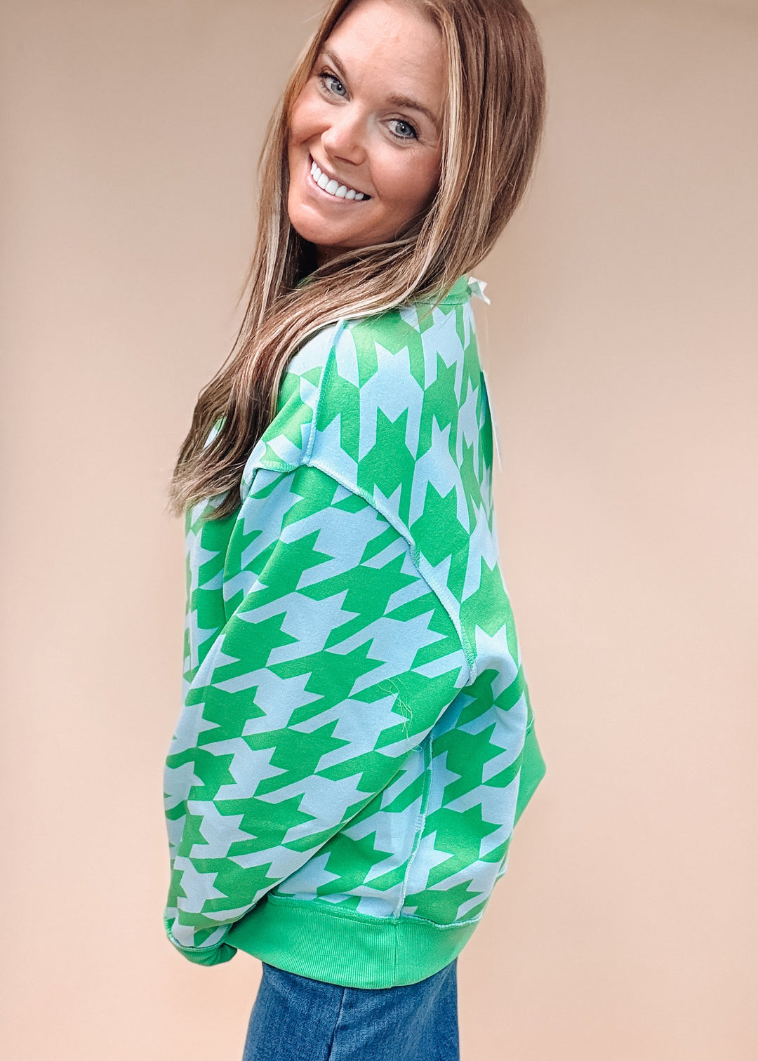 A woman wearing a green and light blue houndstooth pattern sweatshirt and blue jeans, posing against a solid beige background.