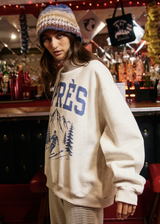 A woman wearing a cream-colored crewneck sweatshirt with a blue graphic print, a striped knit beanie, and striped pants, standing in a warmly lit bar or restaurant setting.