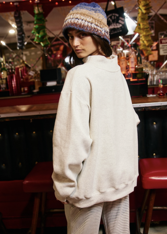A woman in a cozy bar setting wearing an oversized cream-colored sweatshirt, striped pants, and a multicolor knit beanie hat, looking back over her shoulder.