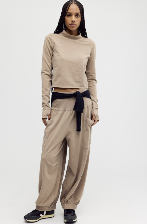 A model wearing a beige long-sleeve crop top and matching beige jogger pants with a dark navy tie belt, paired with black sneakers, against a plain light gray studio background.