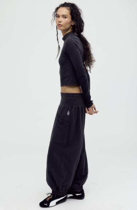 A model wearing a black long-sleeve crop top and matching black wide-leg sweatpants, standing in profile against a plain light gray studio background.