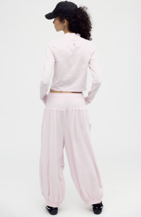 A model from behind wearing a light pink two-piece lounge set consisting of a cropped long-sleeve top and matching wide-leg harem pants, paired with a black baseball cap and black shoes on a white studio background.