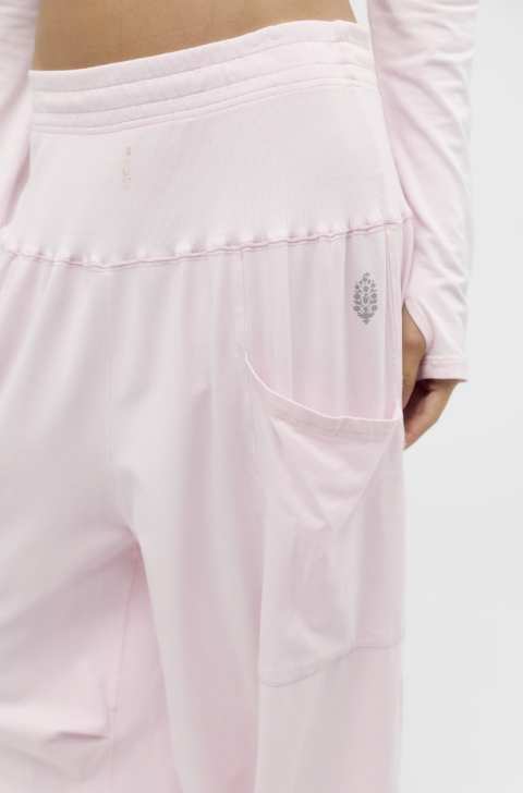 Close-up of a person wearing light pink athletic pants with a side pocket and a small gray embroidered logo, paired with a matching long-sleeve top against a plain light background.