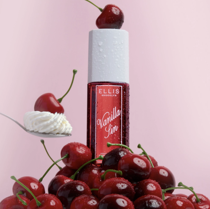 Ellis London Vanilla Sin perfume bottle with a red label and white cap, surrounded by fresh cherries and whipped cream on a pink background.