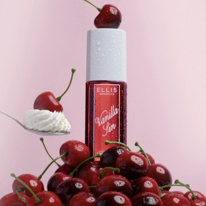 Ellis London Vanilla Sin perfume bottle with a red label and white cap, surrounded by fresh cherries and whipped cream on a pink background.