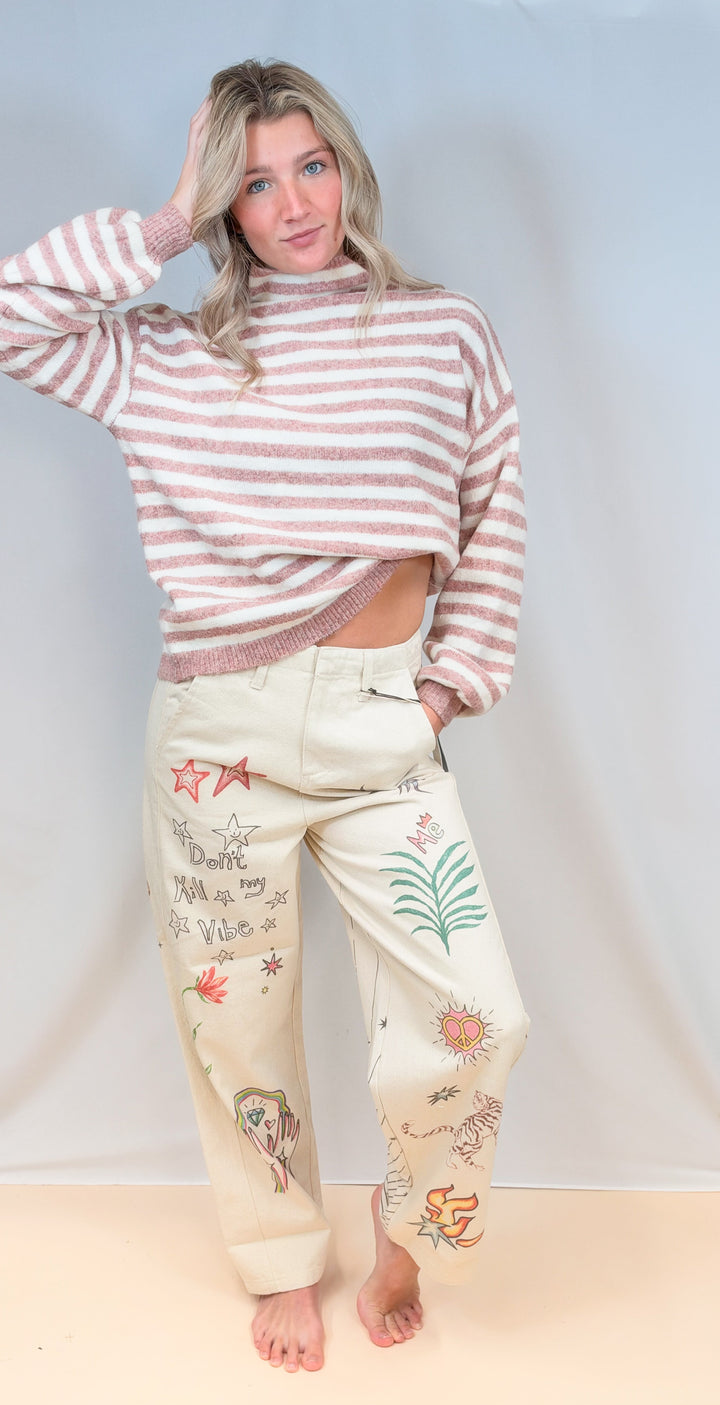Woman wearing pink and cream stripe mock neck sweater with beige barrel pants with doodles on plain background