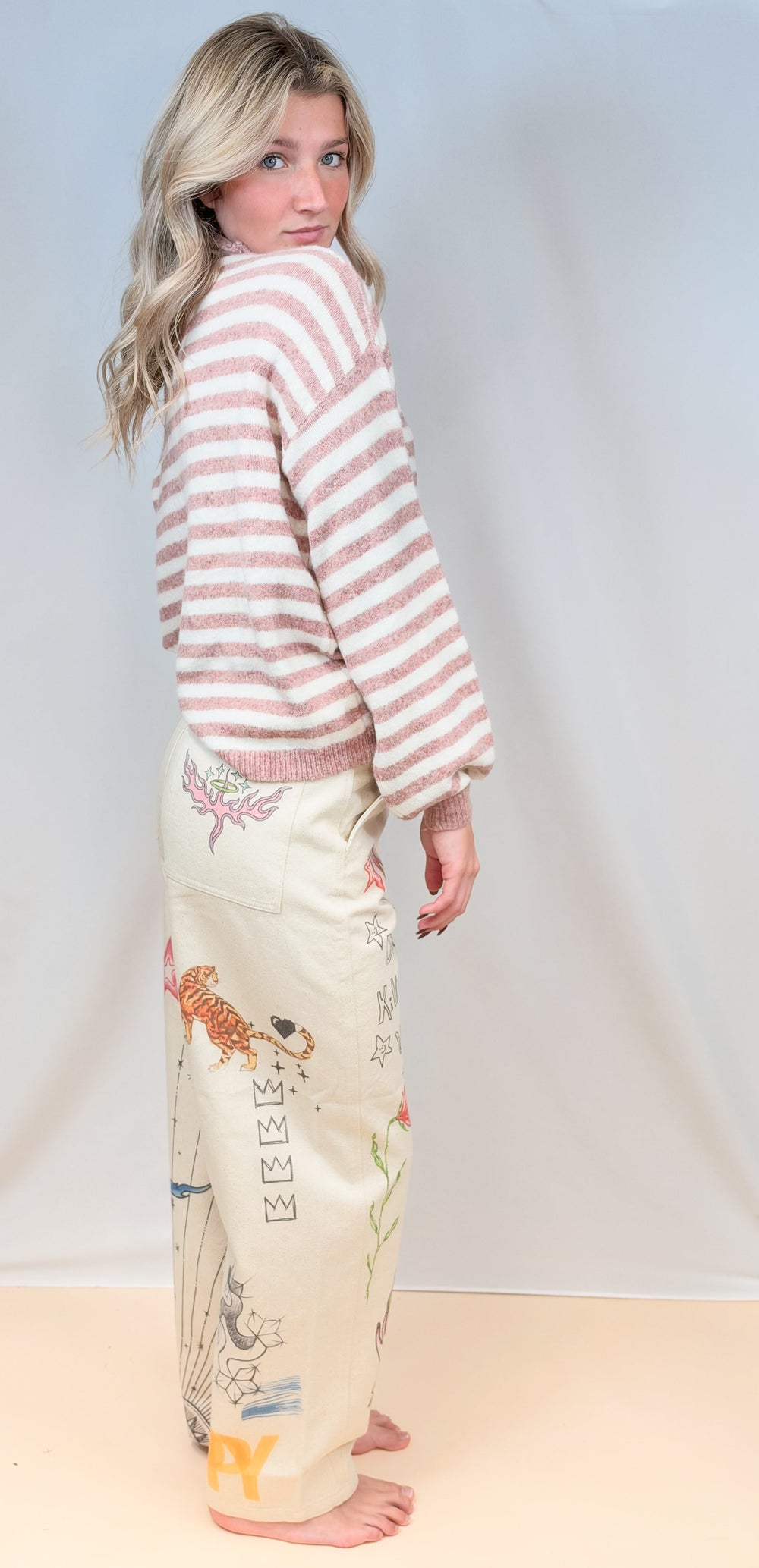 Woman shown from side view wearing pink and cream stripe mock neck sweater with beige barrel pants with doodles on plain background