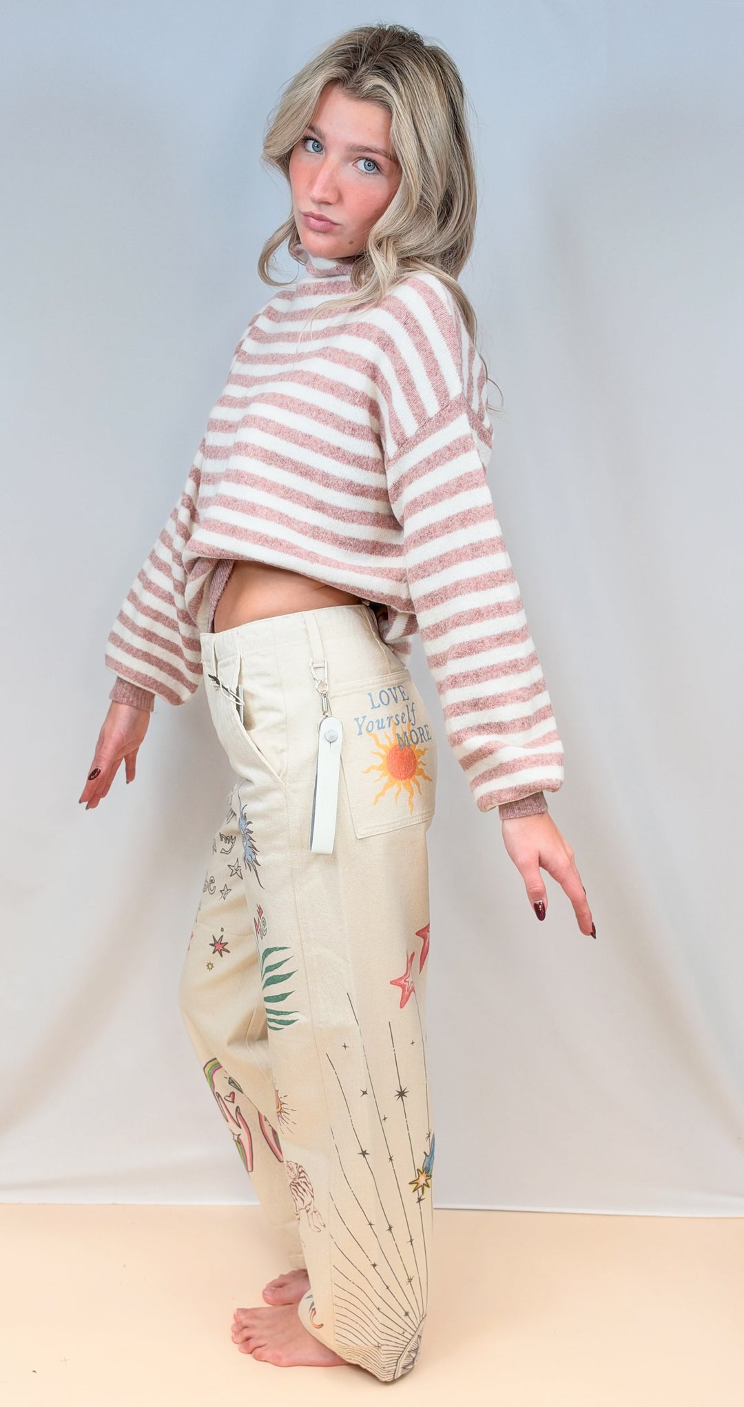 Woman wearing pink and cream stripe mock neck sweater with beige barrel pants with doodles on plain background