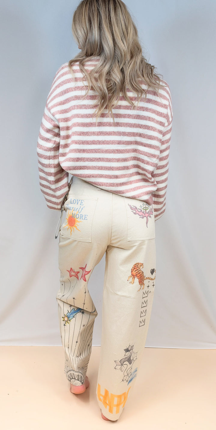 Woman shown from back wearing pink and cream stripe sweater with beige barrel pants with doodles on plain background