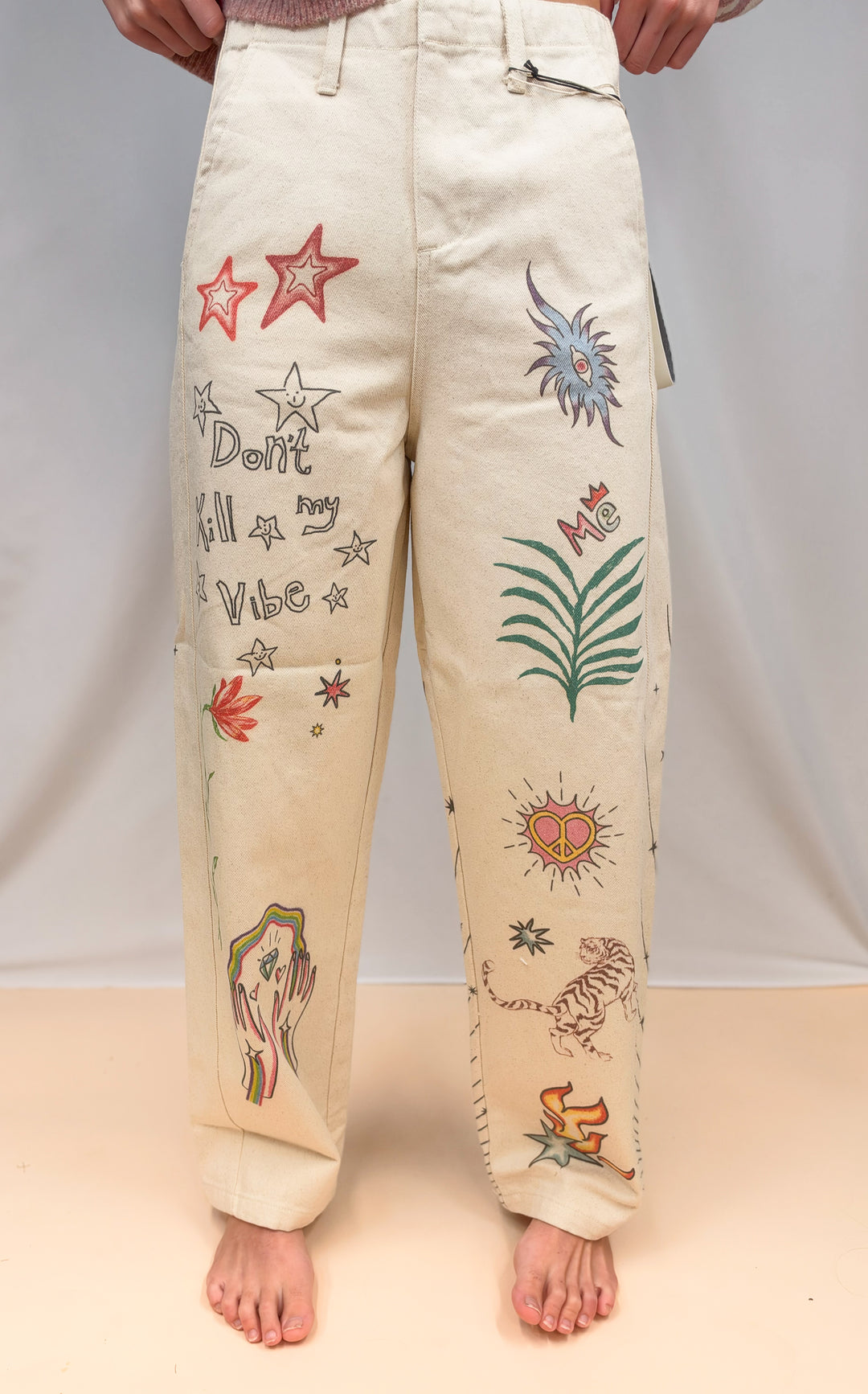 Woman shown from waist down wearing beige barrel pants with doodles on them on plain background