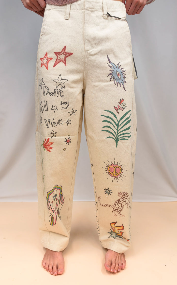 Woman shown from waist down wearing beige barrel pants with doodles on them on plain background