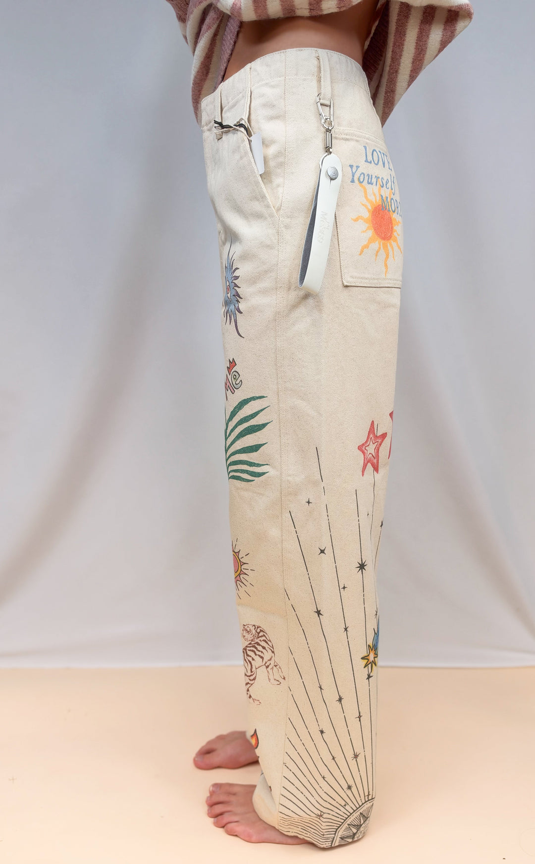 Woman shown from side view wearing beige barrel pants with doodles on plain background