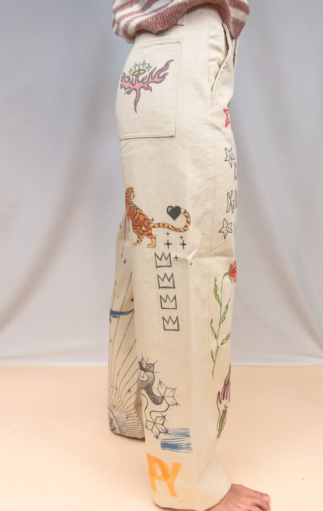 Woman shown from side view wearing beige barrel pants with doodles on plain background