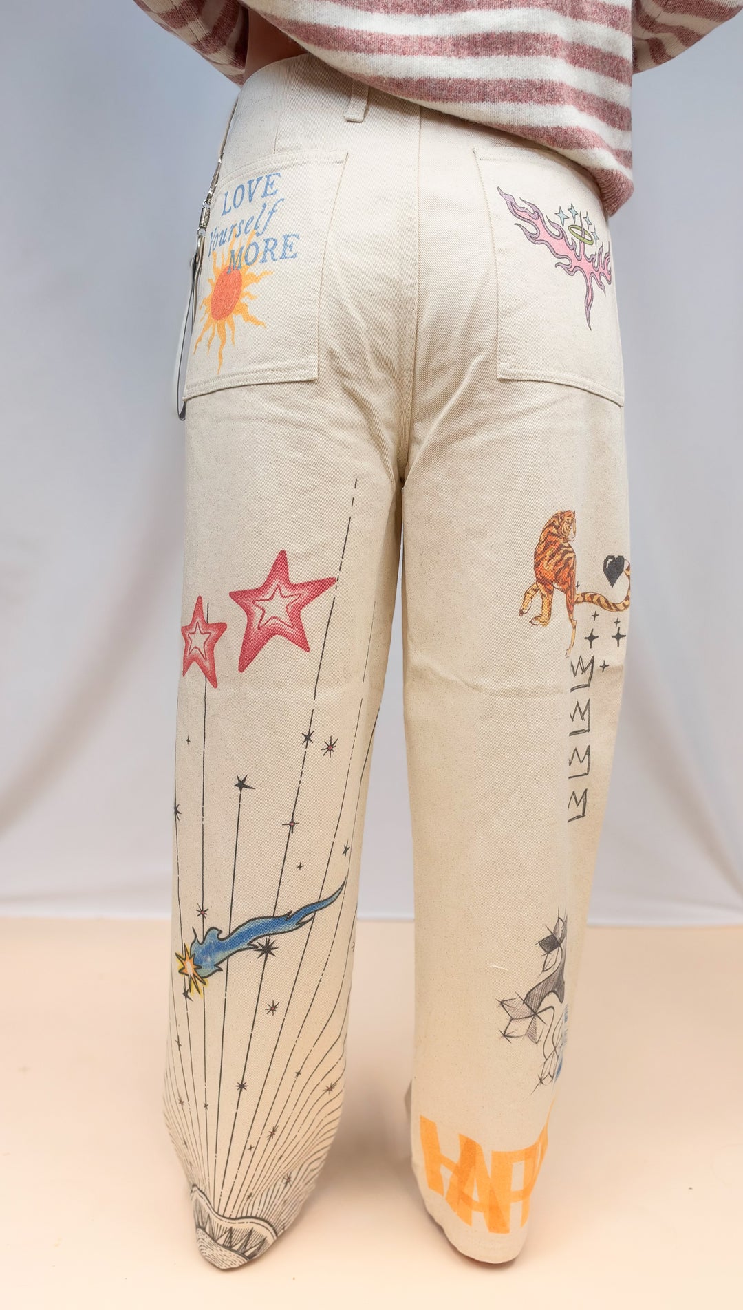 Woman shown from back view wearing beige barrel pants with doodles on plain background