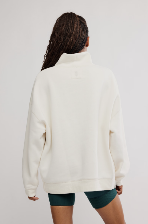 A woman viewed from behind wearing an oversized off-white quarter-zip sweatshirt and dark teal fitted shorts against a plain light gray studio background.