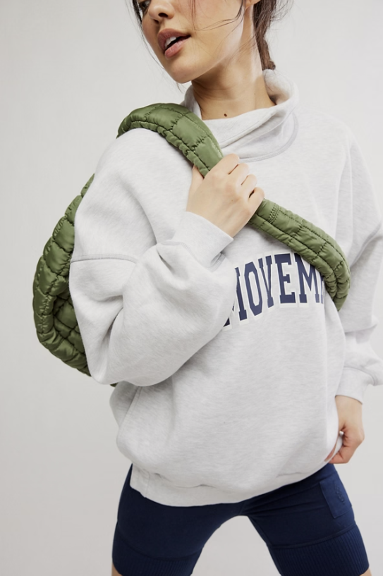 A model wearing a light gray sweatshirt with navy blue collegiate lettering and carrying an olive green quilted shoulder bag.