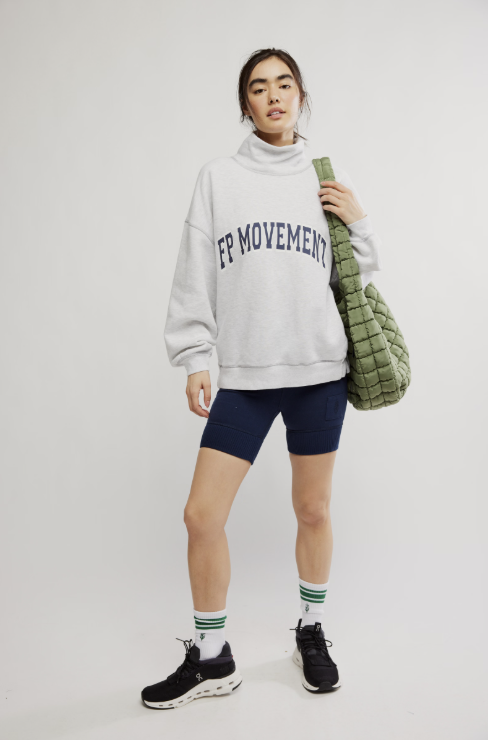 A model stands against a light gray studio background wearing an oversized light gray turtleneck sweatshirt with &