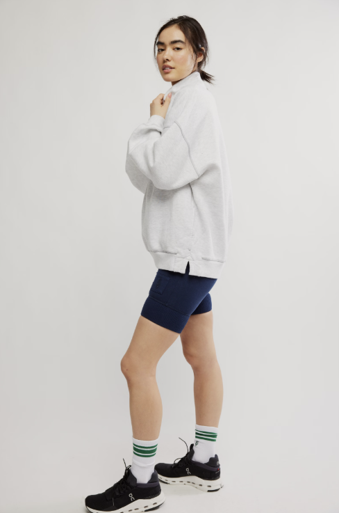 A model wearing a light gray oversized sweatshirt, navy blue fitted biker shorts, white crew socks with green stripes, and black sneakers, posed against a solid light gray studio background.
