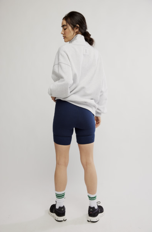 A model is shown from the back wearing a white oversized sweatshirt, navy blue bike shorts, white crew socks with green stripes, and black sneakers, against a plain light gray studio background.