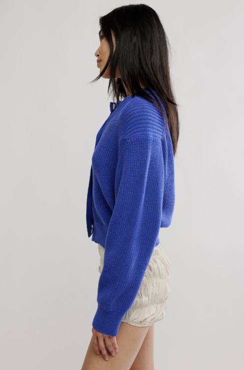 A woman shown in side profile wearing a textured royal blue cropped knit cardigan and cream pleated shorts against a plain light gray studio background.
