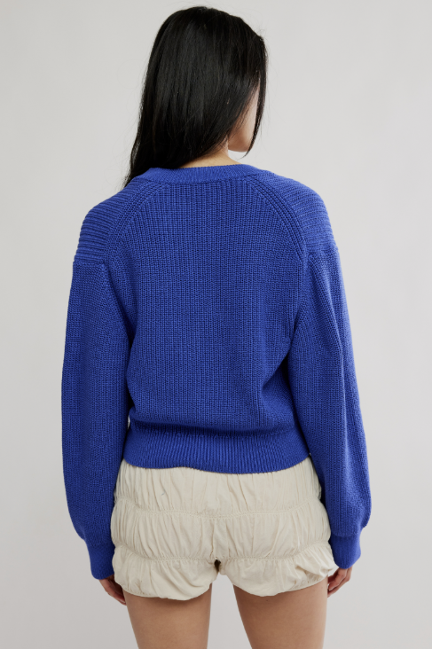 A woman shown from the back wearing a vibrant royal blue ribbed knit sweater and cream-colored ruched shorts against a plain light gray background.