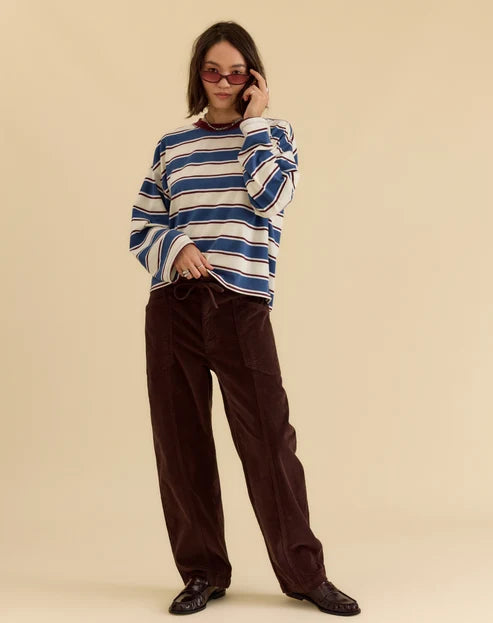 A model wearing a blue, white, and beige striped long-sleeve knit sweater and dark brown corduroy trousers, standing against a solid beige background.