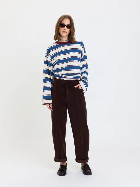 A model wearing a blue and white striped long-sleeve knit top, dark brown wide-leg corduroy trousers, and black loafers, standing against a plain white studio background.