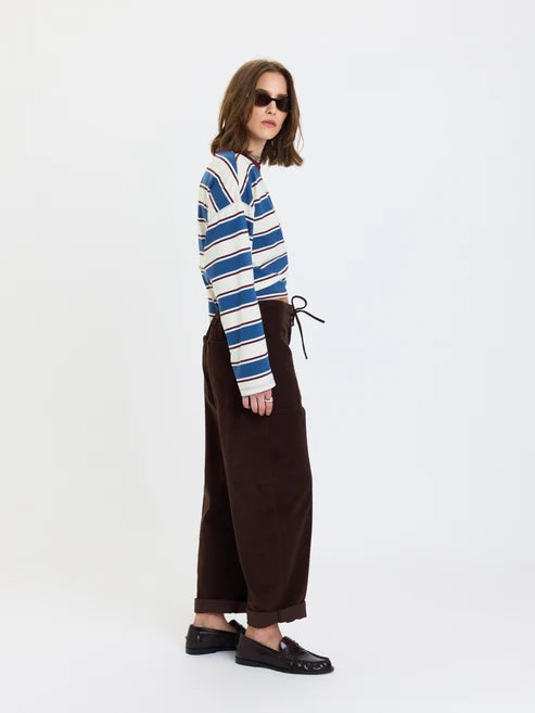 A model is shown from the side wearing a blue, white, and navy striped long-sleeve shirt tucked into dark brown wide-leg trousers, paired with black loafers, against a plain white studio background.