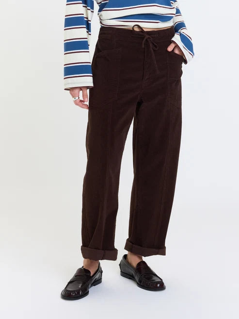 A person wearing dark brown corduroy wide-leg pants with a drawstring waist, paired with a blue, white, and brown striped long-sleeve top and black loafers, shown against a plain white studio background.