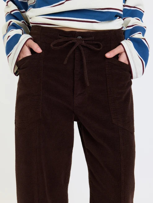 A close-up of a person wearing dark brown corduroy pants with a drawstring waist and side pockets, paired with a blue, white, and burgundy striped long-sleeve top.