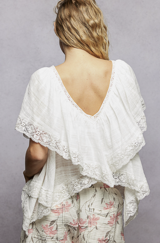 A woman shown from the back wearing a white lace-trimmed draped top with a deep V-neck and a floral print skirt against a gray studio background.