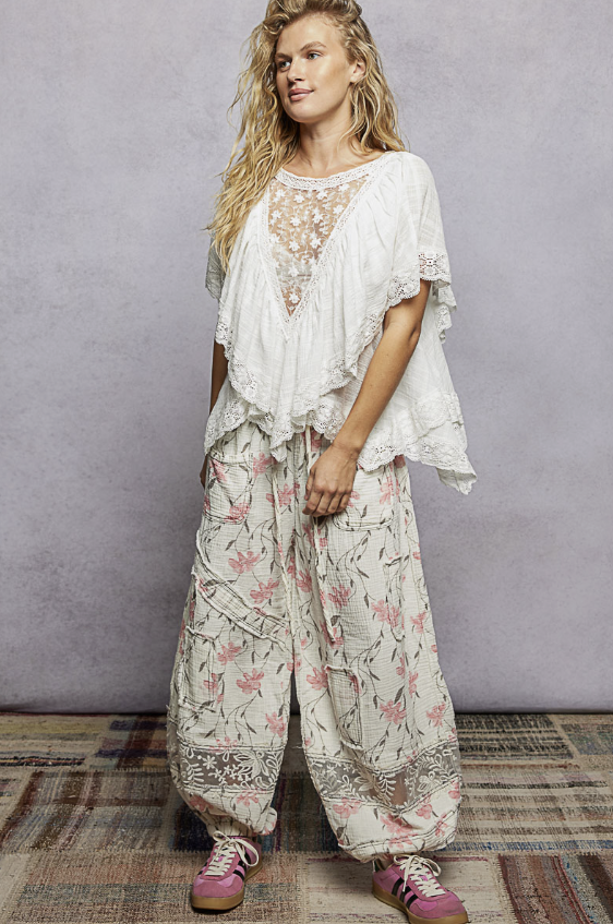 A woman modeling a bohemian-style two-piece outfit featuring a white lace-trimmed tunic top and matching floral print wide-leg pants, paired with pink sneakers, set against a textured gray studio backdrop.