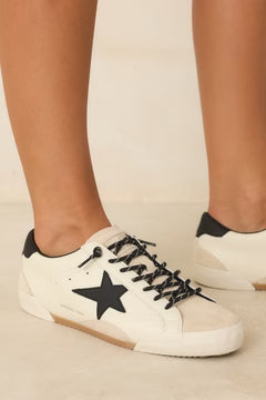White leather low-top sneakers with black star details, black laces, and black heel accents, shown on a model's feet against a light beige background.